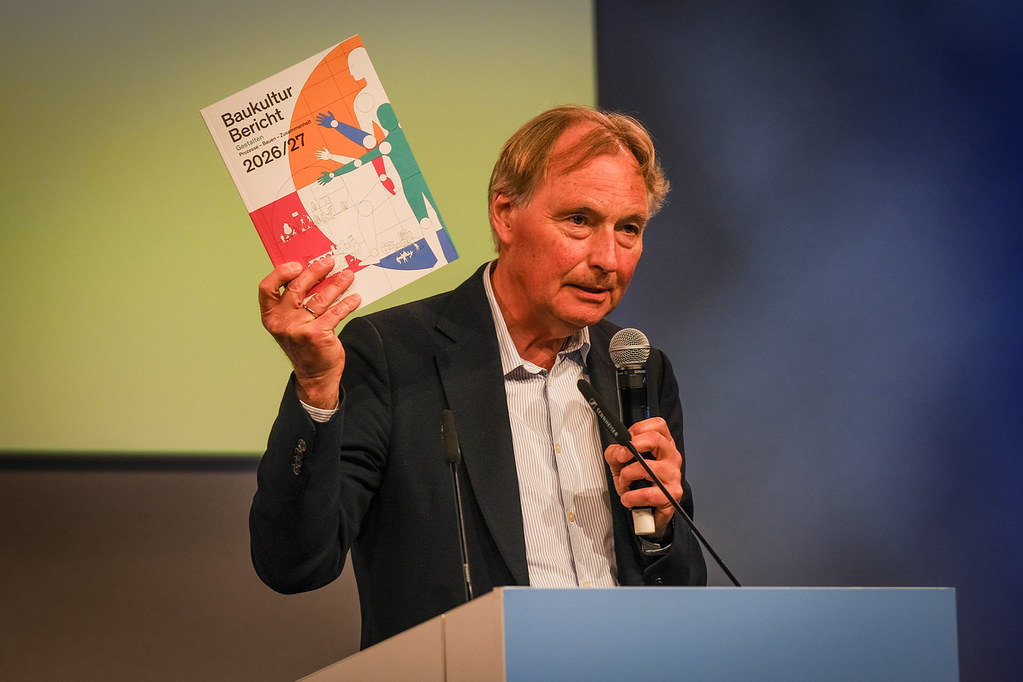 Mann am Rednerpult hält den „Baukultur Bericht 2026/27“ in die Kamera.