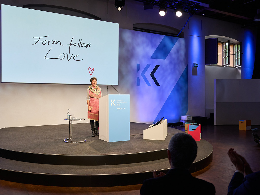 Frau spricht auf einer Bühne vor einer Leinwand mit dem Schriftzug „Form follows Love“.