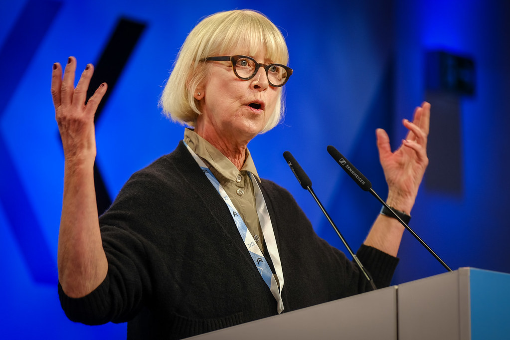 Frau mit Brille und blonden Haaren gestikuliert leidenschaftlich bei einer Rede am Podium.