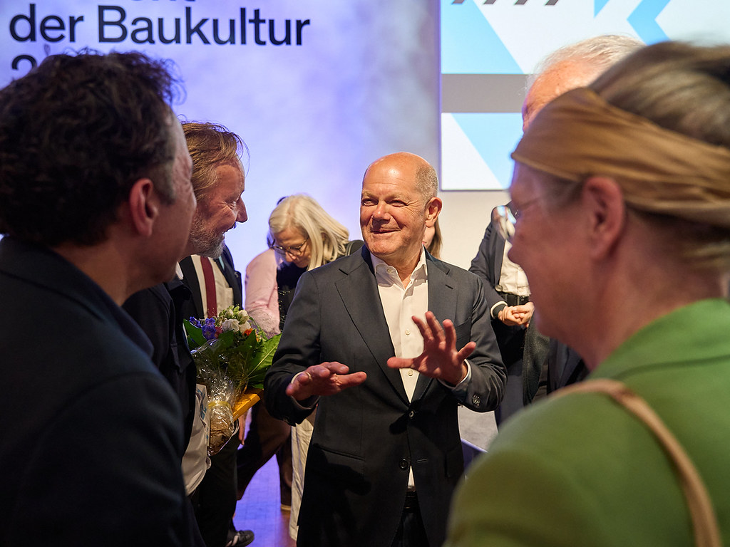 Lachender Mann im Anzug gestikuliert bei einer Veranstaltung zur Baukultur in einer Personengruppe.