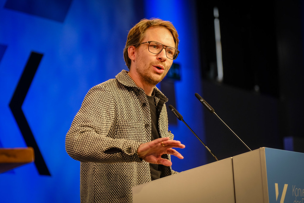 Mann mit Brille und strukturiertem Sakko spricht an einem Rednerpult vor blauem Hintergrund.