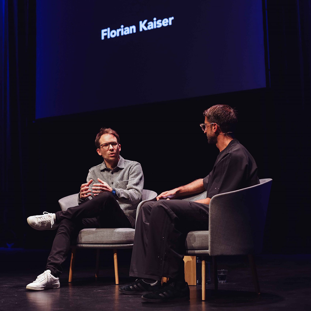 Zwei Männer im Gespräch auf einer Bühne, Podium mit Name Florian Kaiser im Hintergrund.