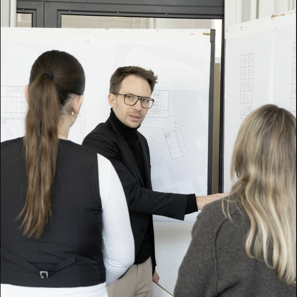 Kolleginnen und Kollege diskutieren vor Bauplänen an einer Whiteboard-/Planwand.