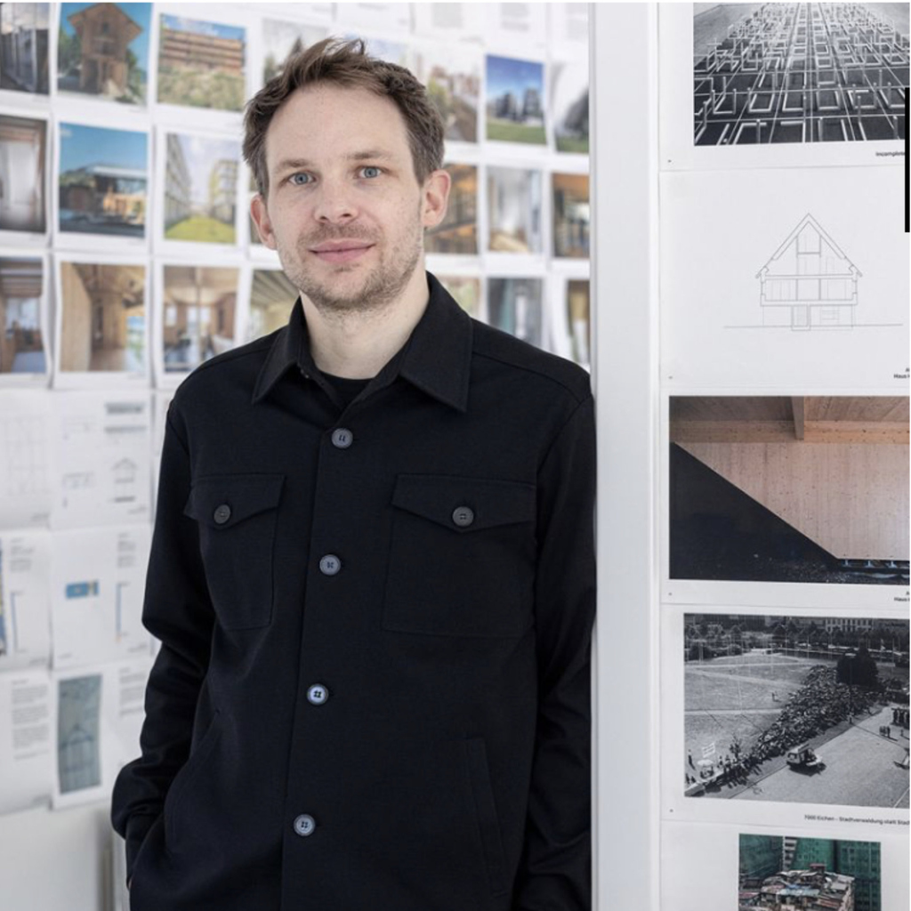 Person steht vor Wand mit Architekturzeichnungen und Fotos, trägt schwarze Jacke.