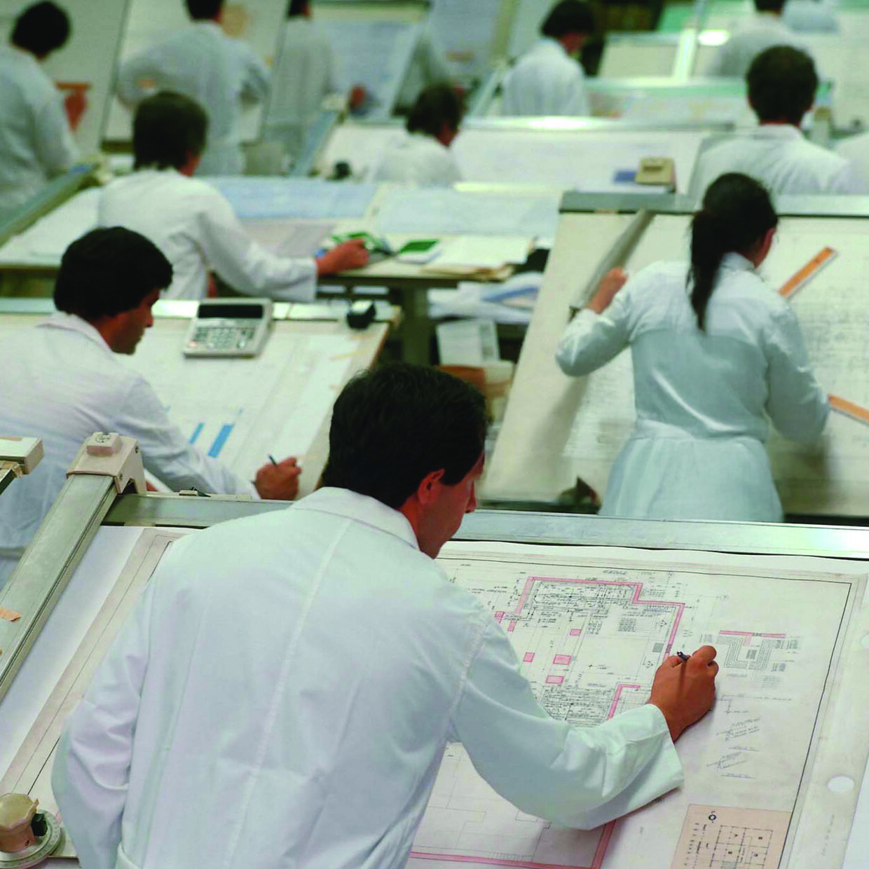 Mehrere Personen in weißen Kitteln arbeiten an technischen Zeichnungen an Zeichentischen.