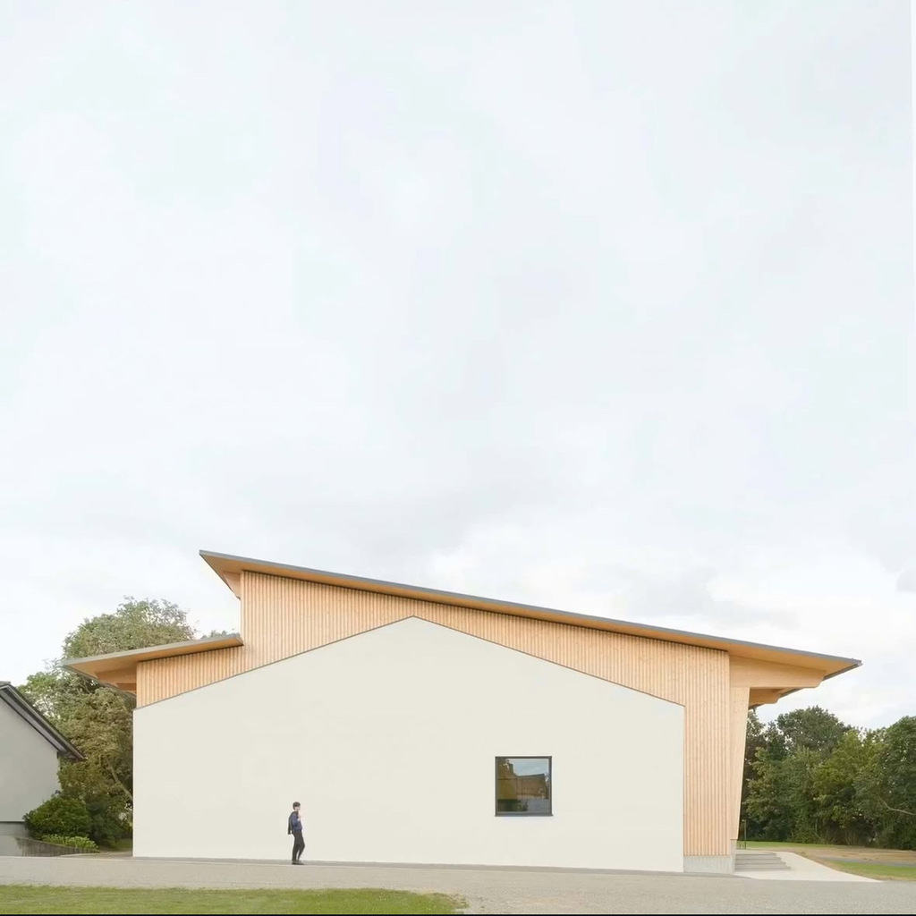 Ein weißes Gebäude mit geneigtem Dach, seitlich Holzelemente, Person steht davor.