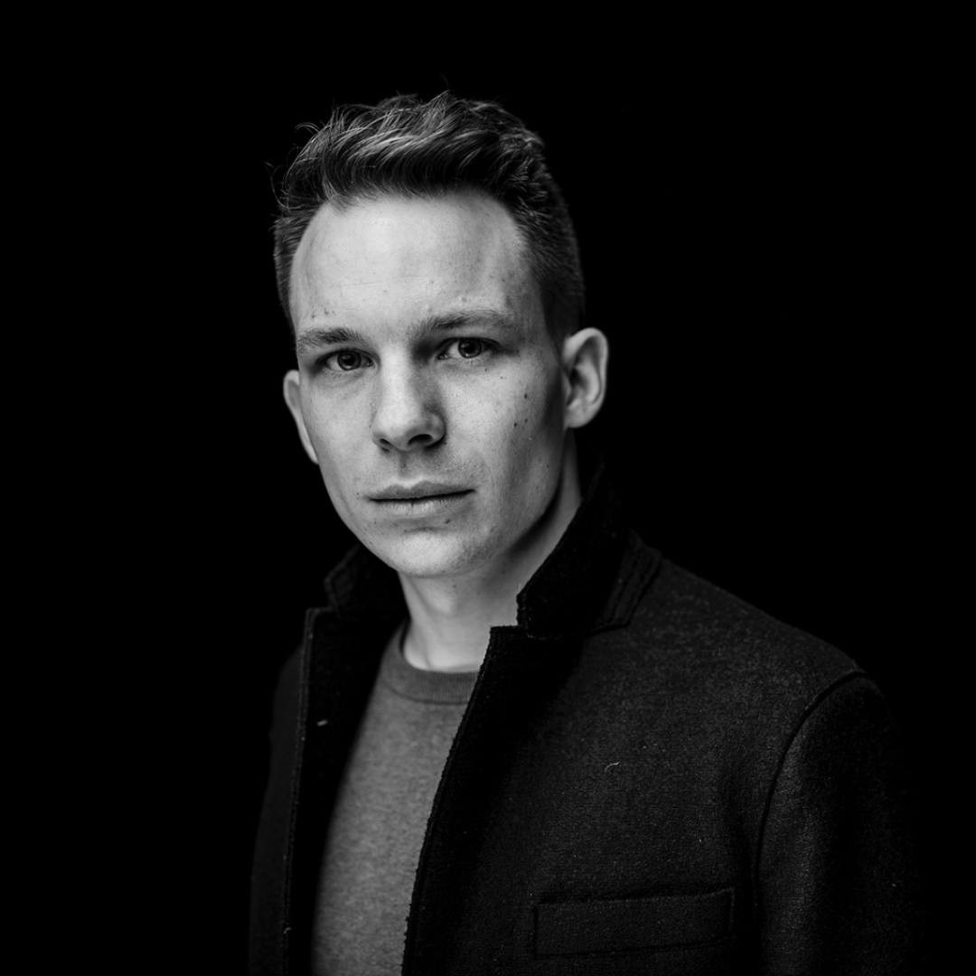 Schwarz-weiß-Porträt eines Mannes mit dunklem Mantel vor schwarzem Hintergrund.