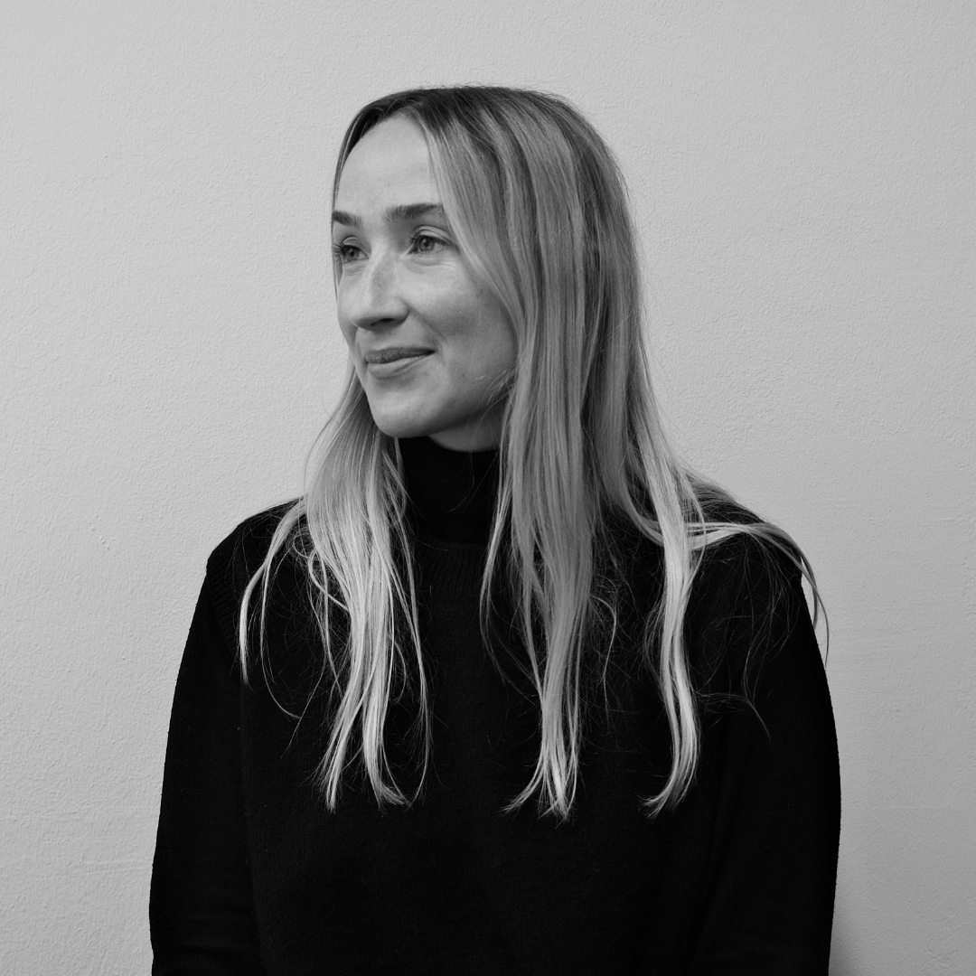 Frau mit langem, blondem Haar und schwarzem Pullover vor einer hellen Wand.