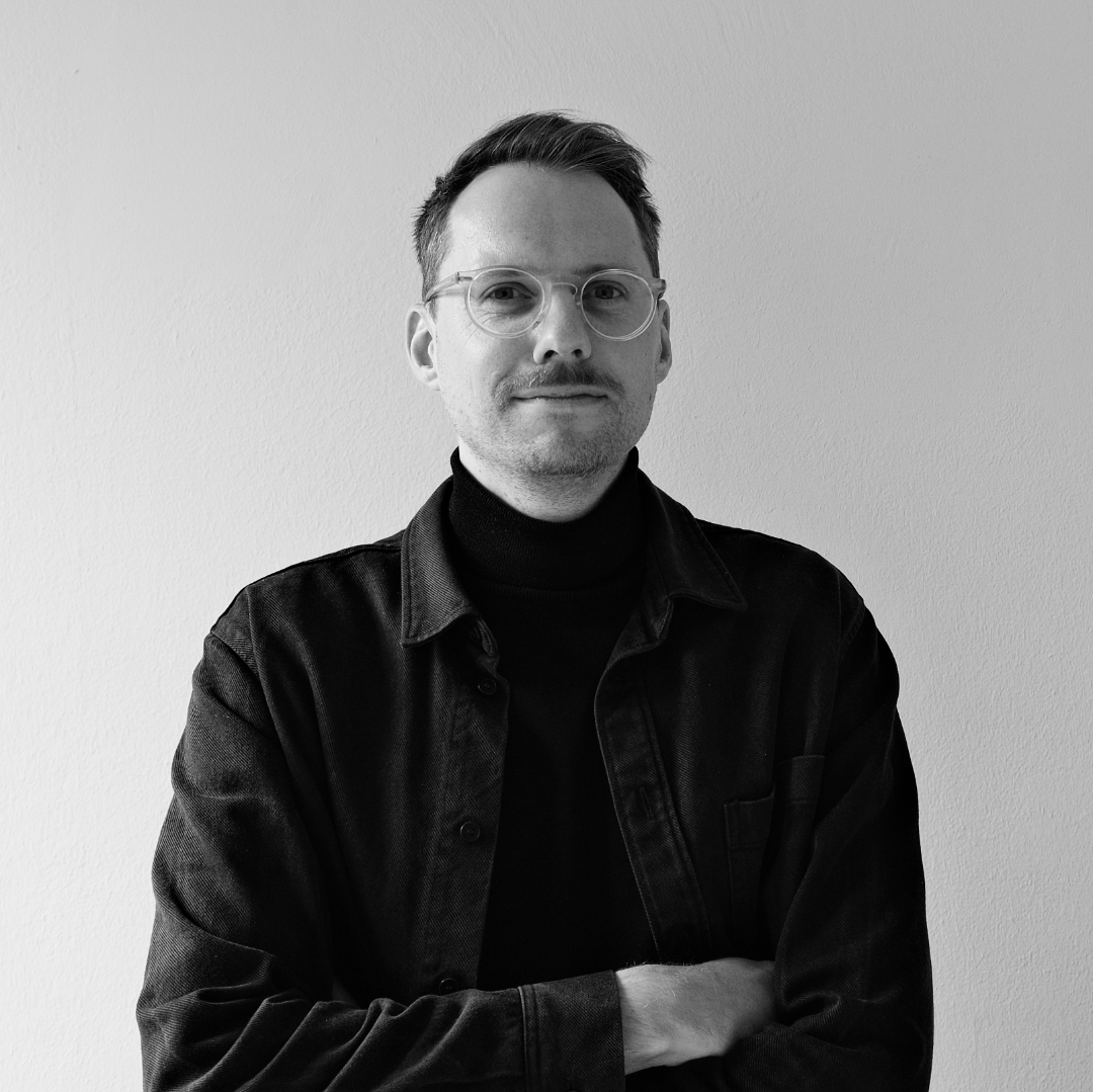 Schwarz-weiß Porträt eines Mannes mit verschränkten Armen vor weißer Wand.